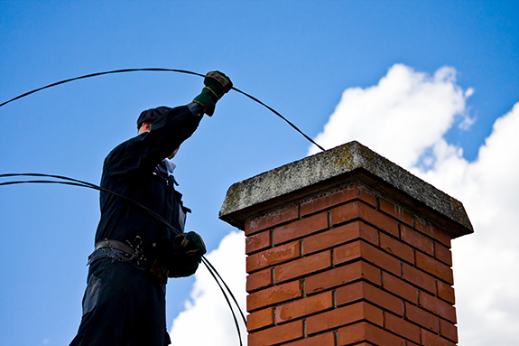 A man in Michigan cleans a chimney with a brush while silhouetted against blue sky and clouds.