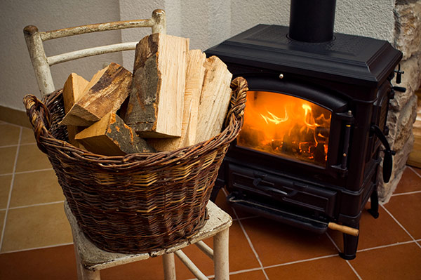 A fire burns brightly in a wood stove and a basket with additional firewood sits on a nearby chair.