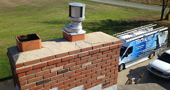 A cracked chimney crown viewed from a rooftop with a Doctor Flue truck parked in the driveway behind it, ready to make chimney masonry repair.
