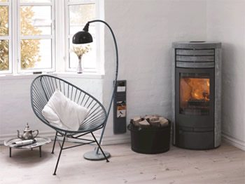 Tall gray Wittus wood stove with a glass door beside a reading chair